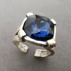 May include: A silver-toned ring featuring a large, square-cut, deep blue faceted crystal stone set in a four-prong metal mounting. The band has an open design, allowing for adjustment, and displays a matte, textured finish on the metal surface.