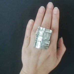 May include: A silver ring with a textured, geometric design. The ring is large and chunky, and it is worn on the index finger.