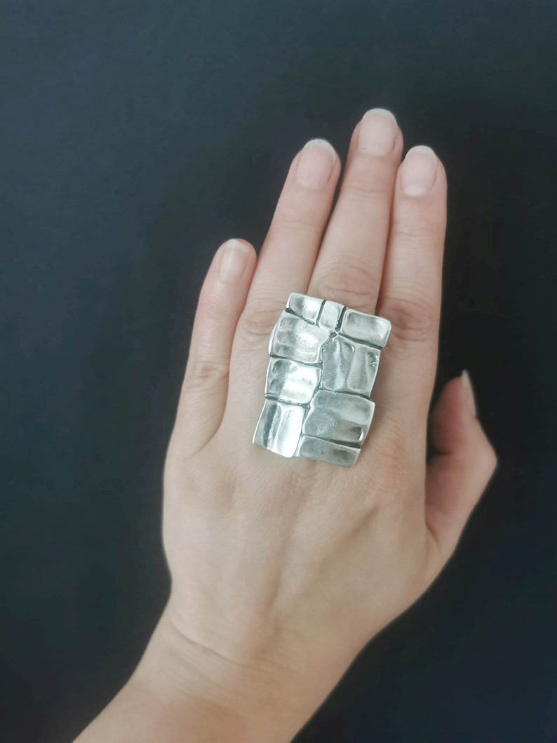 May include: A silver ring with a textured, geometric design. The ring is large and chunky, and it is worn on the index finger.