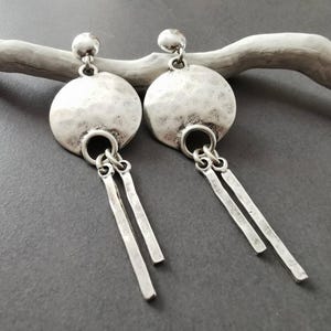 Könnte beinhalten: Ein Paar silberfarbene Ohrringe. Jeder Ohrring hat eine gehämmerten, kreisförmigen Scheibe mit einem hängenden Paar rechteckiger Stäbe. Die Ohrringe sind an einem kleinen, runden Stecker befestigt. Die Ohrringe werden auf einem hellen, strukturierten Ast vor grauem Hintergrund präsentiert.