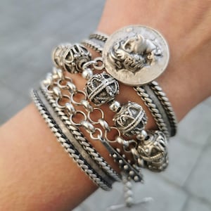 Può includere: Una collezione di braccialetti d'argento con disegni intricati. I braccialetti includono stili intrecciati, a catena e con perline, insieme a un ciondolo a forma di moneta con un rilievo dettagliato. I gioielli sono esposti su un polso.