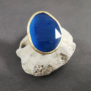 Peut inclure: Une bague ovale avec une grande pierre pr&eacute;cieuse bleue. La pierre est sertie dans une monture dor&eacute;e. La bague repose sur une surface textur&eacute;e blanc cass&eacute;. La pierre a une facette triangulaire blanche.