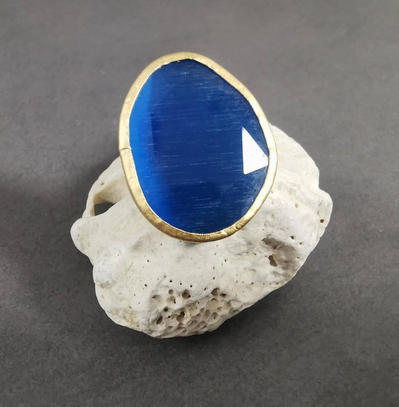 Peut inclure: Une bague ovale avec une grande pierre pr&eacute;cieuse bleue. La pierre est sertie dans une monture dor&eacute;e. La bague repose sur une surface textur&eacute;e blanc cass&eacute;. La pierre a une facette triangulaire blanche.