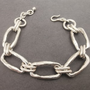 Può includere: Un braccialetto d'argento con grandi maglie ovali e spesse. Le maglie sono collegate da maglie rotonde più piccole.