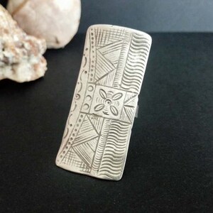 Op de afbeelding: Een zilveren ring met een ingewikkeld geometrisch ontwerp. De ring is rechthoekig en heeft een verhoogd, getextureerd oppervlak.