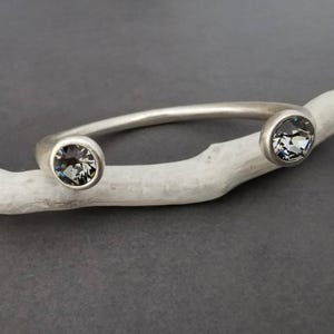 May include: A silver-toned open cuff bracelet featuring a matte finish and two circular settings at each end. Each setting contains a faceted, dark gray crystal that reflects light, creating a contrast against the smooth, curved metal band.