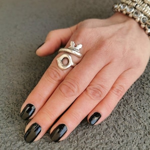 Pode incluir: Um anel prateado com um design de chave e uma abertura circular. O anel é usado numa mão com verniz de unhas preto e uma pulseira de prata. O anel em forma de chave adiciona um toque único à joia.