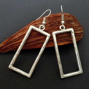 May include: A pair of silver-toned earrings featuring open rectangular frames with a hammered, textured finish. Each earring is suspended from a simple silver-toned hook wire, showcasing a clean, geometric design with a metallic sheen.