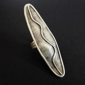 May include: A silver ring with a wavy design on the face. The ring is oval shaped and has a textured finish.