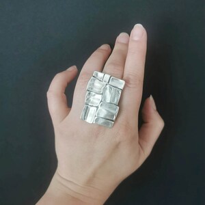 May include: A silver ring with a textured, geometric design. The ring is worn on a person's finger.