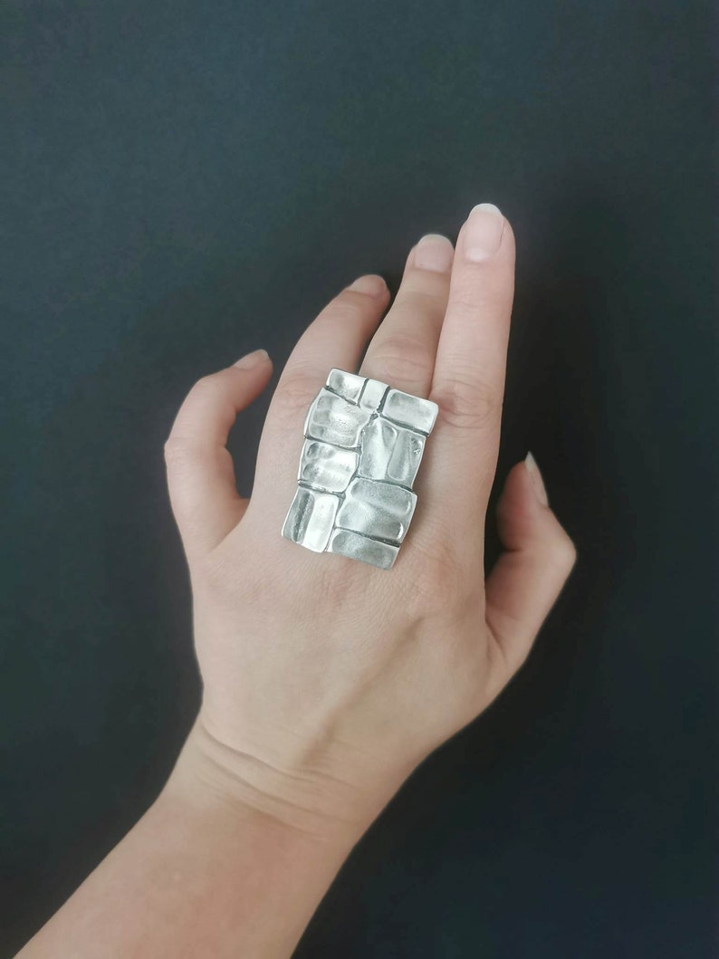 May include: A silver ring with a textured, geometric design. The ring is worn on a person's finger.