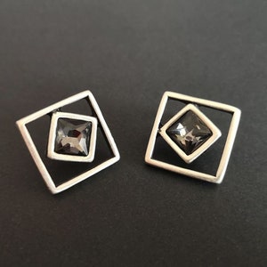 May include: A pair of silver earrings with a square frame and a dark gray crystal in the center.