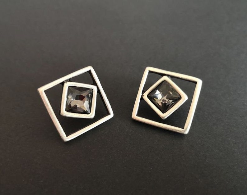 May include: A pair of silver earrings with a square frame and a dark gray crystal in the center.