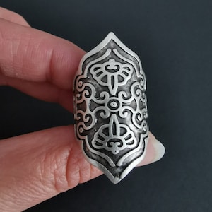 Könnte beinhalten: Ein silberner Ring mit einem komplizierten, verzierten Design. Der Ring ist breit und hat eine spitze Oberseite.
