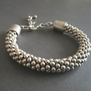 May include: Silver bracelet with a beaded design. The bracelet is made up of small, round beads in a braided pattern. It has a clasp closure and an adjustable chain. The bracelet is displayed on a grey surface.