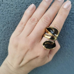 May include: A gold-toned ring with two dark, leaf-shaped accents. The ring has a unique, layered design, with a band of gold and a dark, glossy finish on the leaf-like elements. The ring is worn on a hand with manicured nails.