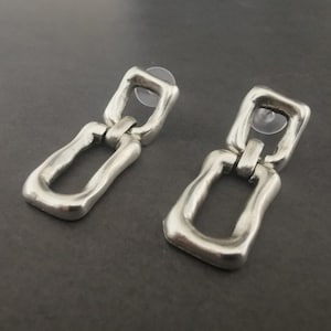 Op de afbeelding: Een paar zilveren oorbellen met een gekoppeld kettingontwerp. Elke oorbel heeft twee rechthoekige schakels met een licht getextureerd oppervlak.