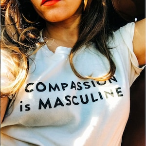 Può includere: Una maglietta bianca con la scritta "COMPASSION is MASCULINE" stampata in nero.