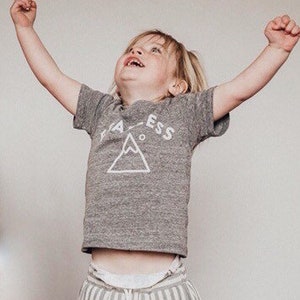 Può includere: Un bambino piccolo che indossa una maglietta grigia con una grafica di montagna bianca e il testo "Fearless" stampato su di essa. Il bambino indossa pantaloni a righe e ha le braccia alzate in aria.