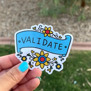 May include: A sticker with a blue banner that reads "VALIDATE" in white letters. The banner is surrounded by yellow and red flowers with green leaves. The sticker is held against a blurred green and brown background.