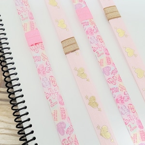 May include: Four pink ribbon bookmarks with gold hearts and arrows. The ribbons have a pink and white pattern with the words "True Love" and "Kiss Me" printed on them.