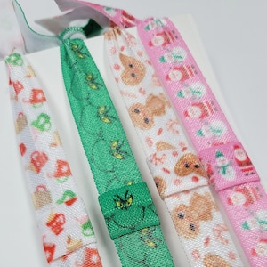 May include: Four colorful hair ties with different holiday-themed patterns. The hair ties are green with bees, white with gingerbread men, white with red teacups, and pink with Santa Claus.