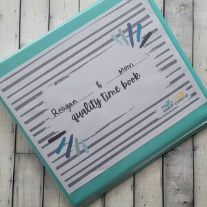 May include: A teal binder with a white label that says "Reagan & Mom quality time book" with a gray and white striped background. The label has a blue and gray paint splatter design. The binder has a "mostly under control" sticker on the side.