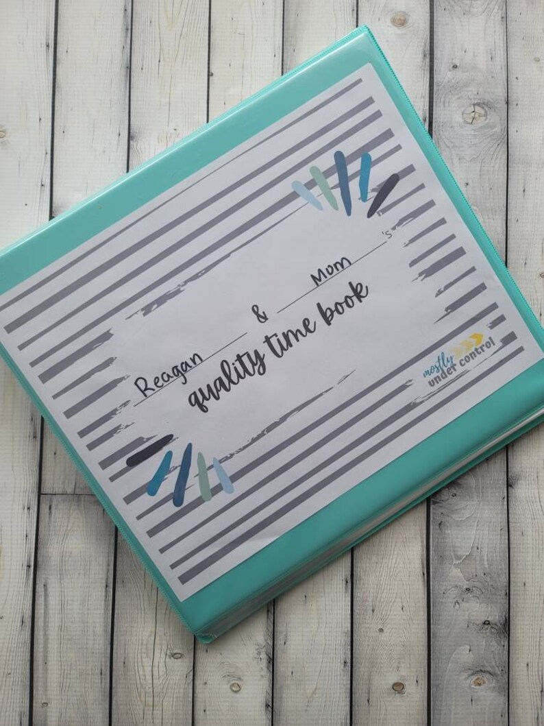May include: A teal binder with a white label that says "Reagan & Mom quality time book" with a gray and white striped background. The label has a blue and gray paint splatter design. The binder has a "mostly under control" sticker on the side.
