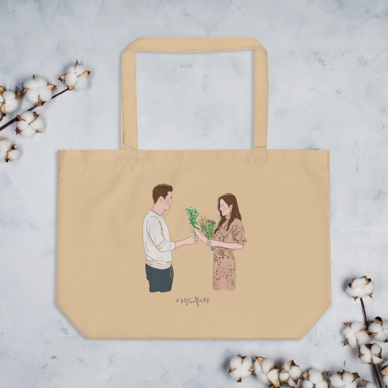 Op de afbeelding: Een beige tote bag met een geprinte illustratie van een stel dat bloemen uitwisselt. De tekst "사랑하는 당신에게" is onder de illustratie gedrukt.