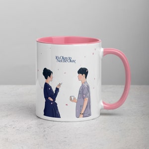 以下が含まれることがあります： ピンクの縁取りと取っ手のある白いセラミックマグカップ。マグカップには、立って互いに見つめ合っている2人の人物のグラフィックが描かれています。マグカップには「It's Okay to Not Be Okay」という文字が印刷されています。