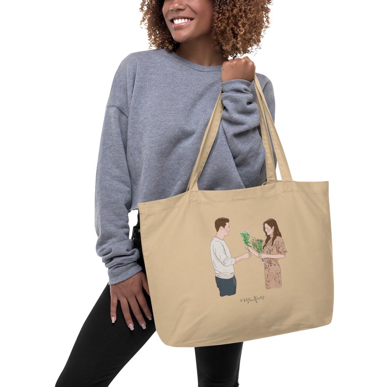 Op de afbeelding: Een beige tote bag met een afbeelding van een man en een vrouw die bloemen vasthouden. De tekst "사랑의 불시착" staat op de tas gedrukt.
