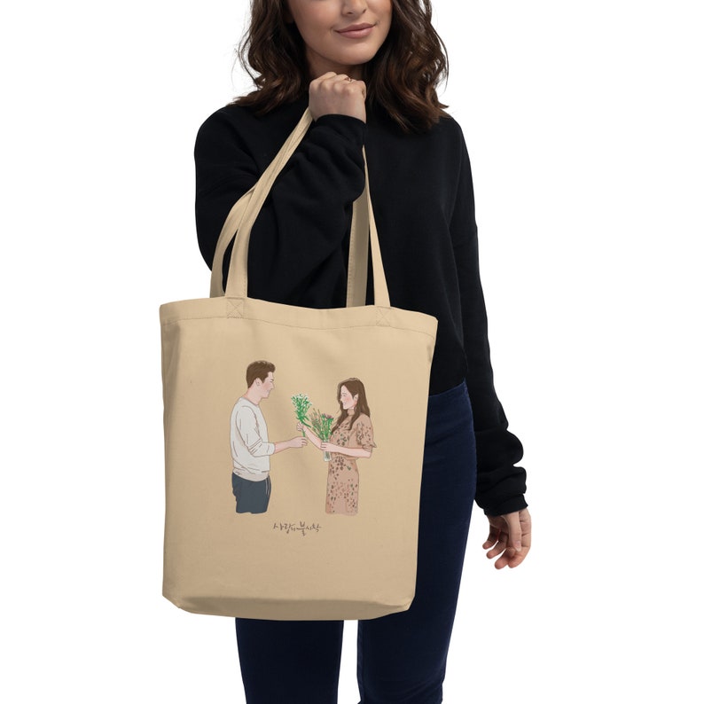 Op de afbeelding: Een beige tote bag met een afbeelding van een stel dat bloemen vasthoudt. De tekst "사랑은 영원히" staat onder de afbeelding.
