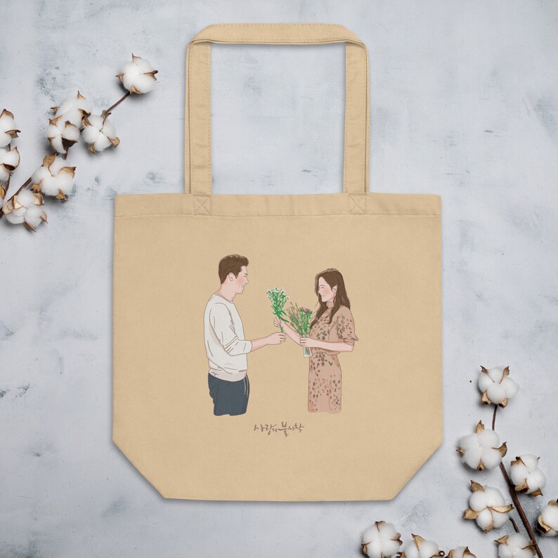 Op de afbeelding: Een beige tote bag met een geprinte illustratie van een stel dat bloemen vasthoudt. De tekst "사랑스러운 날" is onder de illustratie gedrukt.