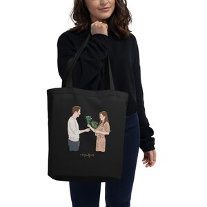 Op de afbeelding: Zwarte tote bag met een afbeelding van een man en een vrouw die bloemen vasthouden. De tekst "사랑의 불시착" is onder de afbeelding gedrukt.