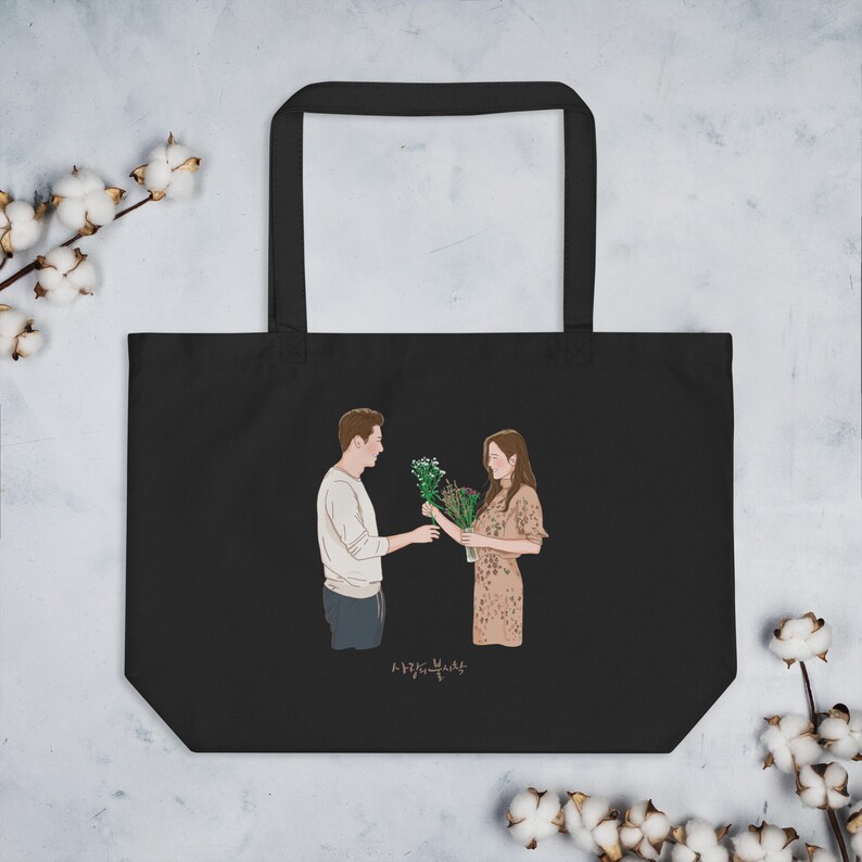 Op de afbeelding: Zwarte tote bag met een afbeelding van een stel dat bloemen vasthoudt. De tekst "사랑의 불시착" staat op de tas gedrukt.