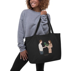 Op de afbeelding: Zwarte tote bag met een afbeelding van een man en een vrouw die boeketten bloemen vasthouden. De tekst "사랑의 불시착" is onder de afbeelding gedrukt.