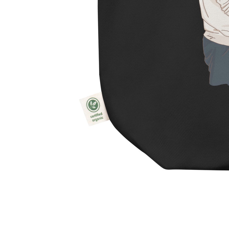 Op de afbeelding: Zwarte tote bag met een witte afbeelding van een persoon. De tas heeft een groen "gecertificeerd biologisch" label.