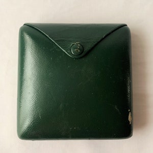 May include: A dark green leather box with a single button closure. The box is slightly worn and has a vintage look.