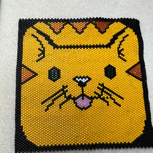 May include: A yellow beaded cat face with green eyes, a pink tongue sticking out, and black whiskers. The cat face is surrounded by a black border.
