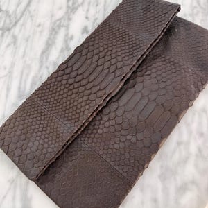 May include: A dark brown rectangular clutch bag featuring a textured snakeskin pattern across the entire exterior surface. The bag has a fold-over flap design with a horizontal seam running across the middle, showcasing a consistent scale texture.