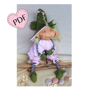 Può includere: Un gnomo di feltro verde con una camicia a righe rosa e bianche e scarpe di feltro verde. Il gnomo tiene una chiave ed è seduto su un pezzo di legno alla deriva.