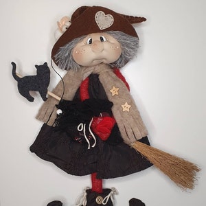 Può includere: Una bambola di strega fatta a mano con un cappello marrone, capelli grigi, un vestito nero e una scopa. La bambola tiene un gatto nero e un guanto rosso.