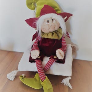 May include: A plush elf doll with a green hat, red and white striped legs, and a red velvet outfit. The elf is sitting on a white pillow with tassels.