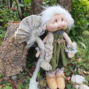 Peut inclure: Une poupée artisanale au design fantaisiste. La poupée a une perruque blanche, une robe verte et des chaussures beiges. Elle tient un accessoire en forme de champignon. La poupée se trouve dans un cadre naturel extérieur.