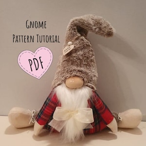 Può includere: Un gnomo di peluche con un cappello di pelliccia marrone, una camicia a quadri rossi e neri, una barba bianca e un fiocco bianco. Lo gnomo è seduto su una superficie bianca. Il cappello ha un'etichetta che dice "Hand Made". L'immagine include il testo "Gnome Pattern Tutorial PDF".