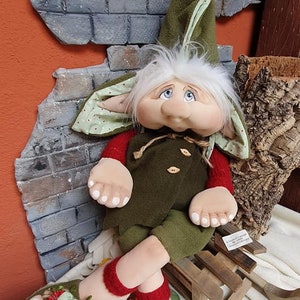 May include: A green and red gnome doll with white hair and a green hat sits on a wooden sledge. The gnome is wearing green trousers and a green jacket with brown buttons. The gnome has a large nose and big blue eyes.