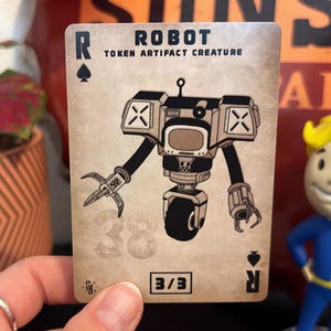 Puede incluir: Una carta de juego rectangular con fondo beige desgastado que presenta una ilustración en blanco y negro de un robot con una sola rueda, manos de garra y un pecho tipo pantalla. El texto dice ROBOT, TOKEN ARTIFACT CREATURE y 3/3.