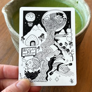 May include: A rectangular white card featuring a black line art illustration of a bowl of ramen pouring noodles into a river, alongside a fish, sushi, a hot air balloon, mountains, and a sign with the Japanese text おいしい and English text YOU GAIN A LIFE.