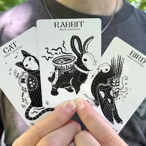 Könnte beinhalten: Drei weiße Spielkarten mit schwarzen Tuschezeichnungen einer Katze, eines Kaninchens und eines Vogels. Jede Karte ist mit dem Tiernamen und dem Text Token Creature beschriftet. Die Katze trägt einen Rucksack, das Kaninchen eine Schale und der Vogel einen Köcher.