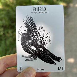May include: A white rectangular game card featuring a black ink illustration of a bird carrying a quiver of arrows on its back. The text BIRD TOKEN CREATURE is printed at the top, with FLYING and 1/1 at the bottom. The bird has intricate patterns.